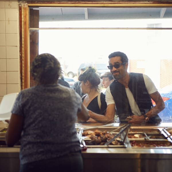 Customers ordering Haitian food from a server behind a counter with several hot food trays.