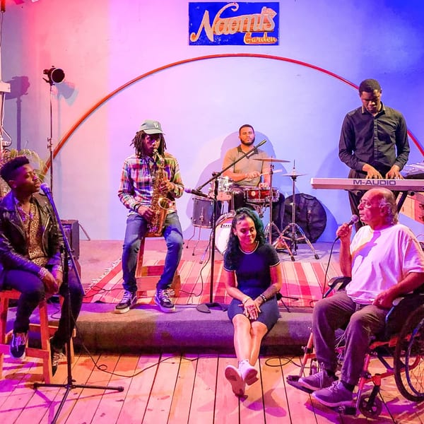 A band performs on stage at Naomi's Garden, with a saxophonist, drummer, keyboardist, and two singers.