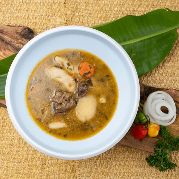 A light blue bowl of Haitian soup with meat, dumplings, and vegetables, garnished with peppers and onions.