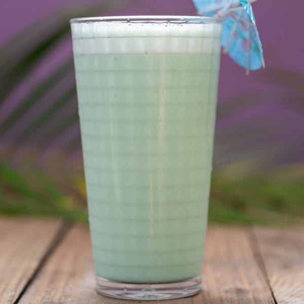 A tall glass of light green Haitian smoothie with a blue umbrella on a wooden table.