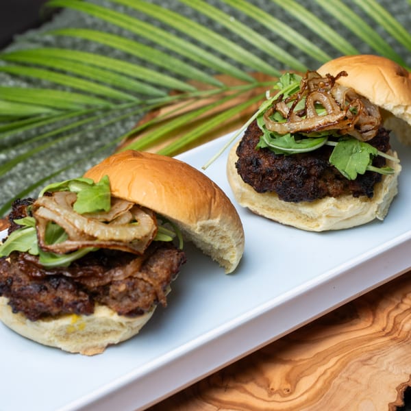 Two Haitian sliders with dark patties, caramelized onions, and arugula on a white plate.