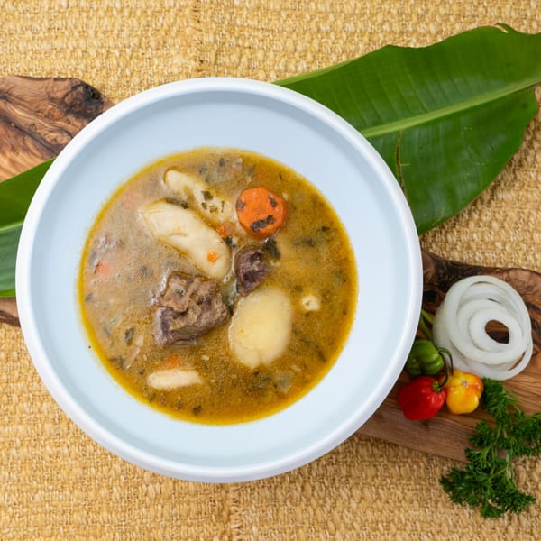 Haitian soup joumou with meat, vegetables, and dumplings in a white bowl, garnished with peppers and onions.
