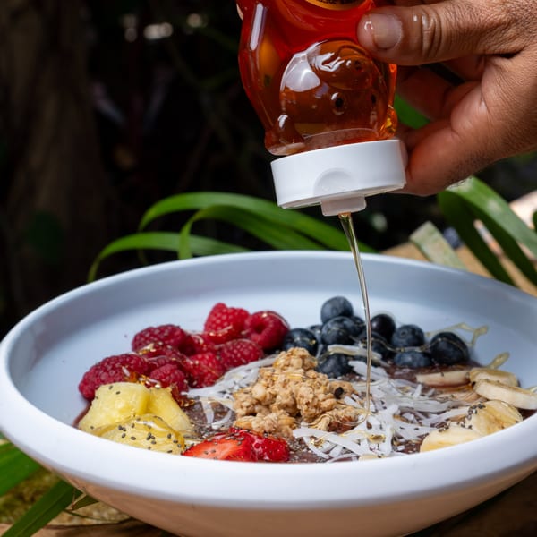 A hand drizzles honey from a bear-shaped bottle onto a colorful fruit and granola bowl.