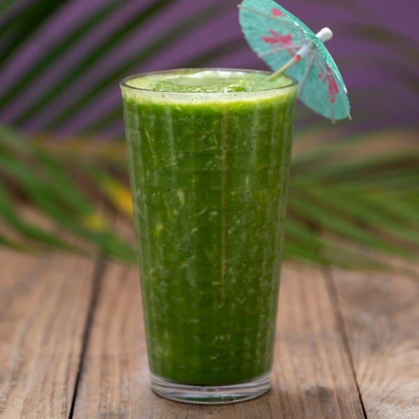 Green smoothie with a small umbrella on a wooden table with palm leaves in the background.