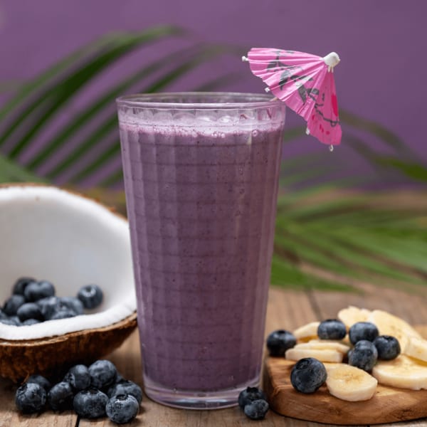 Blueberry smoothie with a pink umbrella, fresh blueberries, and banana slices on a wooden table.