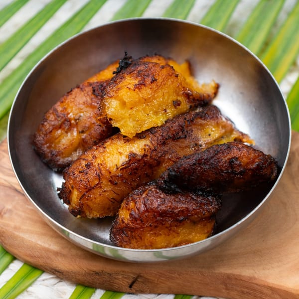 A metal bowl filled with golden-brown fried plantains, served on a wooden board with green leaves.
