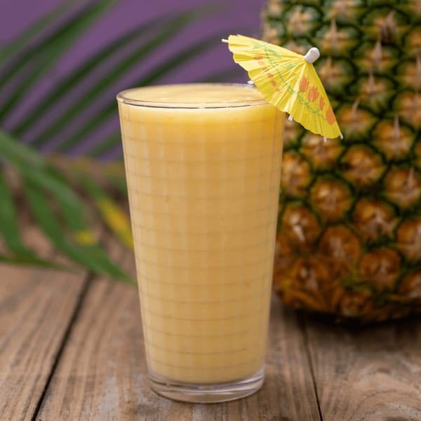 A glass of pineapple smoothie with a yellow umbrella, next to a whole pineapple on a wooden table.