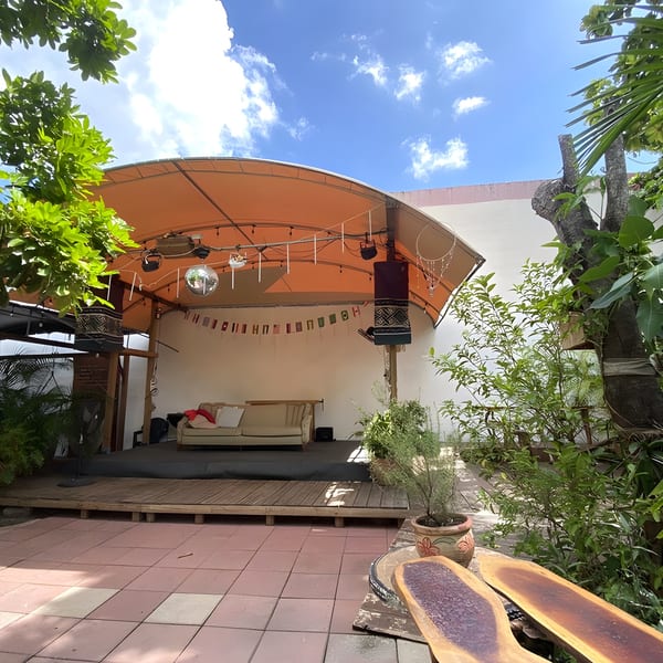 Outdoor lounge area with a stage, sofa, disco ball, and lush greenery under a blue sky.