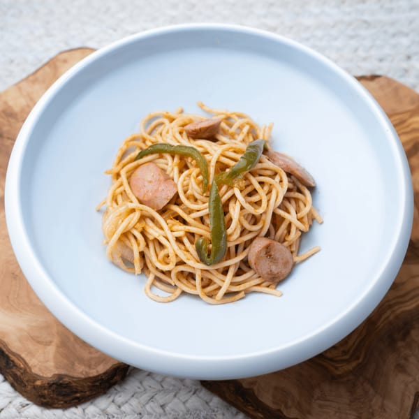 Haitian spaghetti with sliced sausage and green peppers in a light blue bowl on a wooden board.