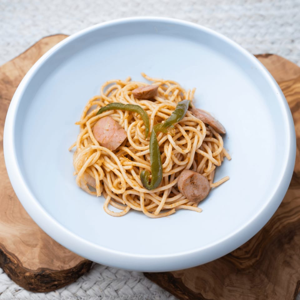 Haitian spaghetti with sliced sausage and green peppers in a light blue bowl on a wooden board.