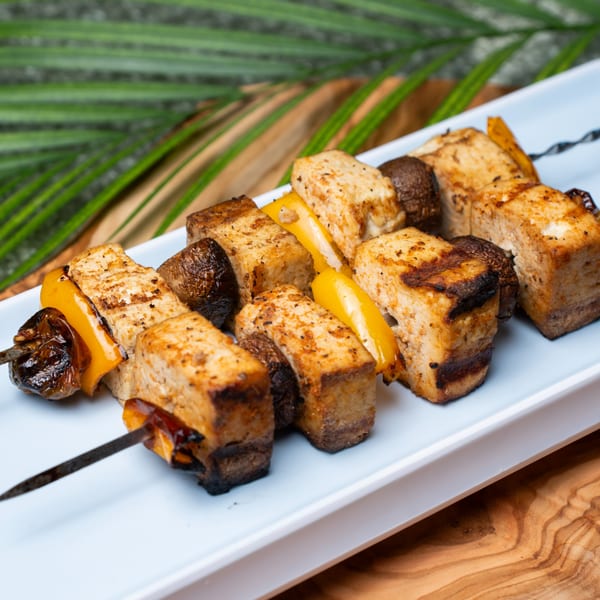 Grilled Haitian tofu, mushroom, and bell pepper skewers on a white plate with palm leaves.