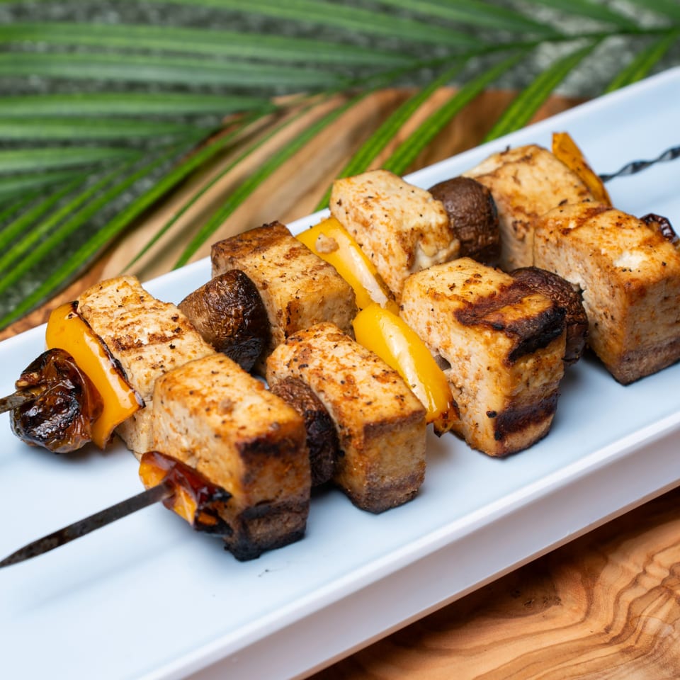 Grilled Haitian tofu, mushroom, and bell pepper skewers on a white plate with palm leaves.