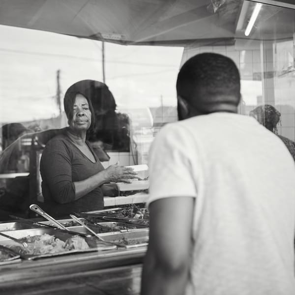A woman serving Haitian food in a takeout container to a customer at Naomi's Garden.
