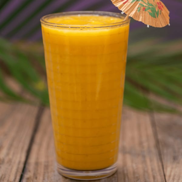 A tall glass of vibrant orange mango smoothie with a small paper umbrella on a wooden table.