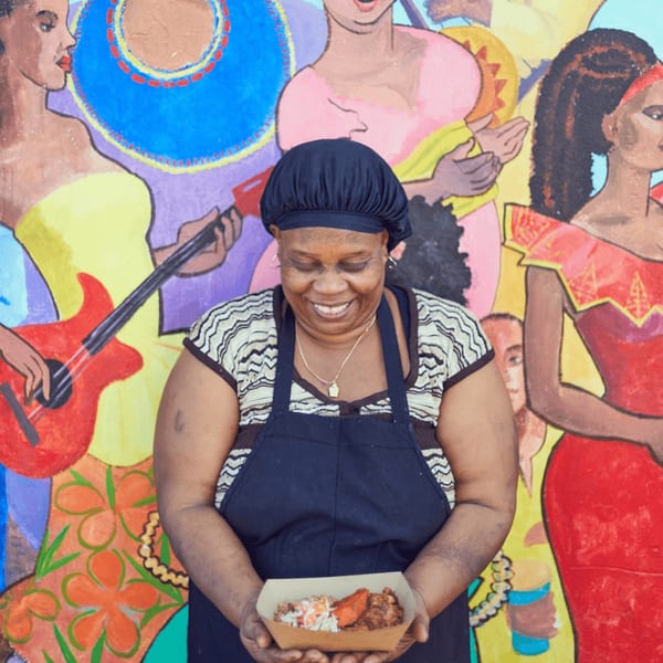 A smiling woman in a black apron holds a Haitian meal in front of a colorful mural.