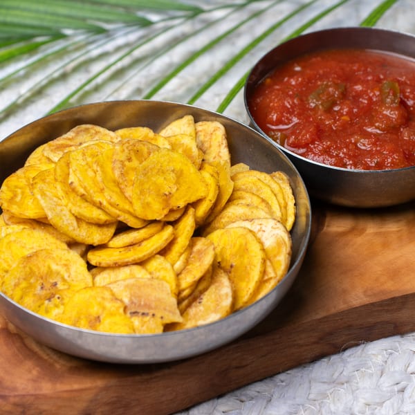 A bowl of crispy Haitian plantain chips next to a bowl of red salsa on a wooden board.