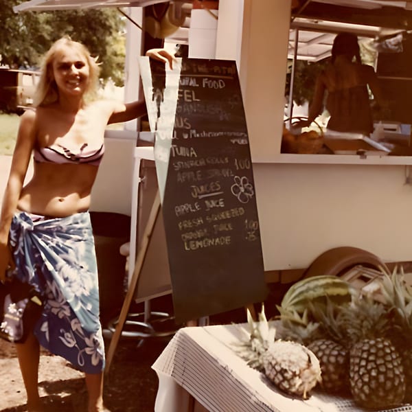 A woman in a bikini and sarong stands next to a food stand with a menu and fresh fruit.