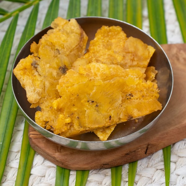 A metal bowl filled with golden-brown Haitian fried plantains, or tostones, on a wooden board.