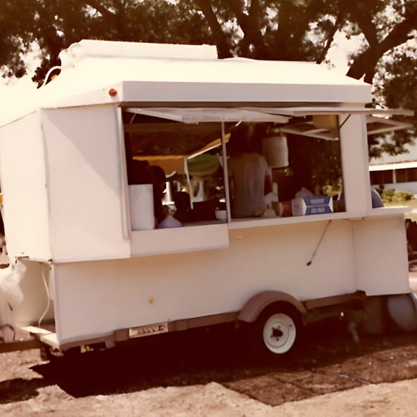 A white food truck with an open serving window, parked outdoors under trees.