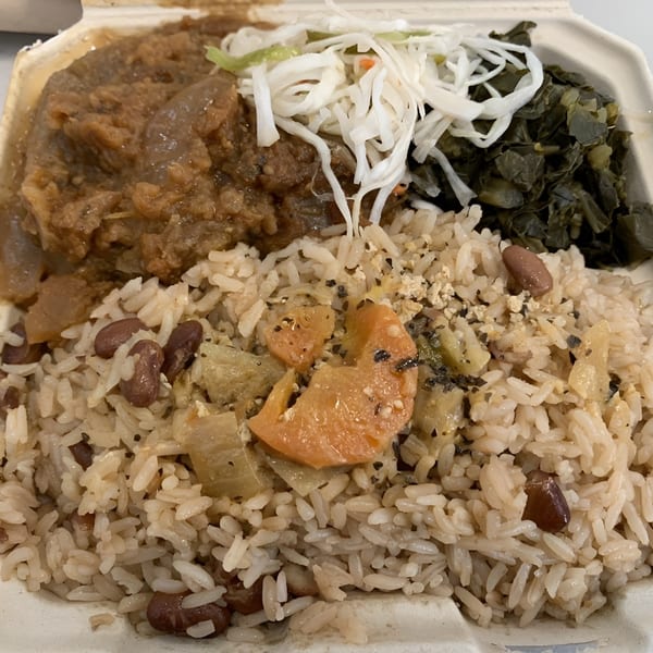 Haitian meal with rice and beans, stewed meat, collard greens, and shredded cabbage in a takeout container.
