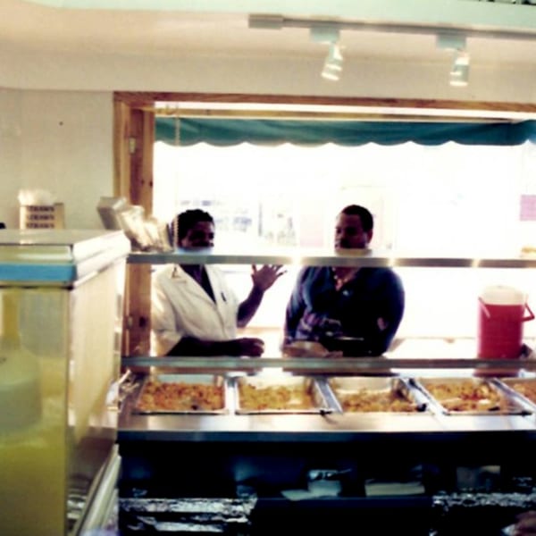 Two men behind a food counter with Haitian dishes in serving trays at Naomi's Garden.