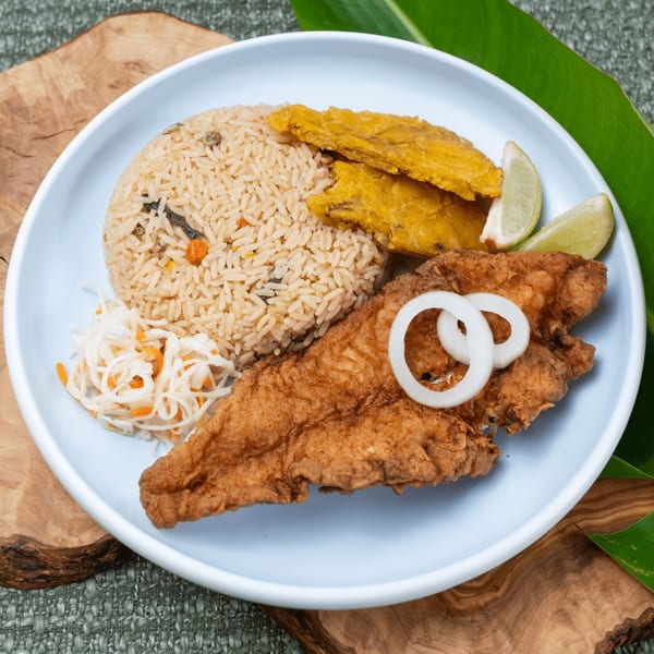 Haitian fried fish with rice, plantains, pikliz, and lime wedges on a light blue plate.