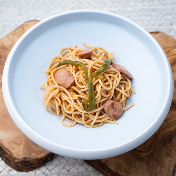 Haitian spaghetti with sliced sausage and green peppers in a light blue bowl on a wooden board.