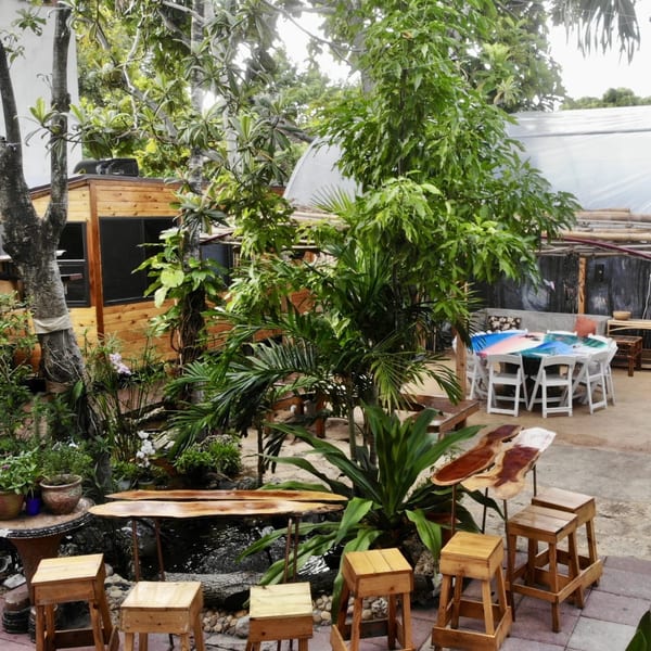 Lush outdoor dining area at Naomi's Garden with wooden stools, tables, and a pond surrounded by tropical plants.