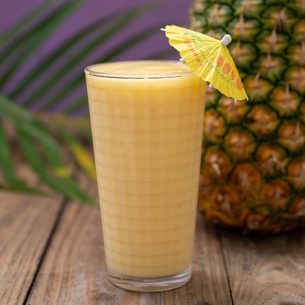 A refreshing pineapple smoothie with a yellow umbrella on a wooden table, with a whole pineapple and palm leaves in the background.