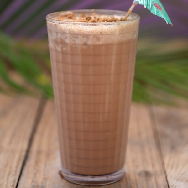 A tall glass of Haitian chocolate drink with foam and a small umbrella on a wooden table.
